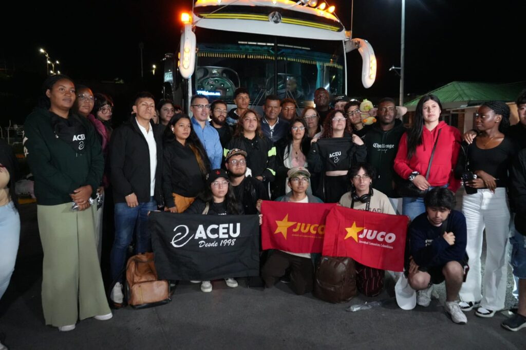 El Gobernador de Nariño, Luis Alfonso Escobar Jaramillo, posa junto a un nutrido grupo de jóvenes líderes en la Terminal de Transporte de Pasto durante la noche, frente a un bus intermunicipal blanco que facilitó su retorno seguro. Los jóvenes sostienen banderas de organizaciones como ACEU y JUCO tras completar el corredor humanitario desde el Cauca.