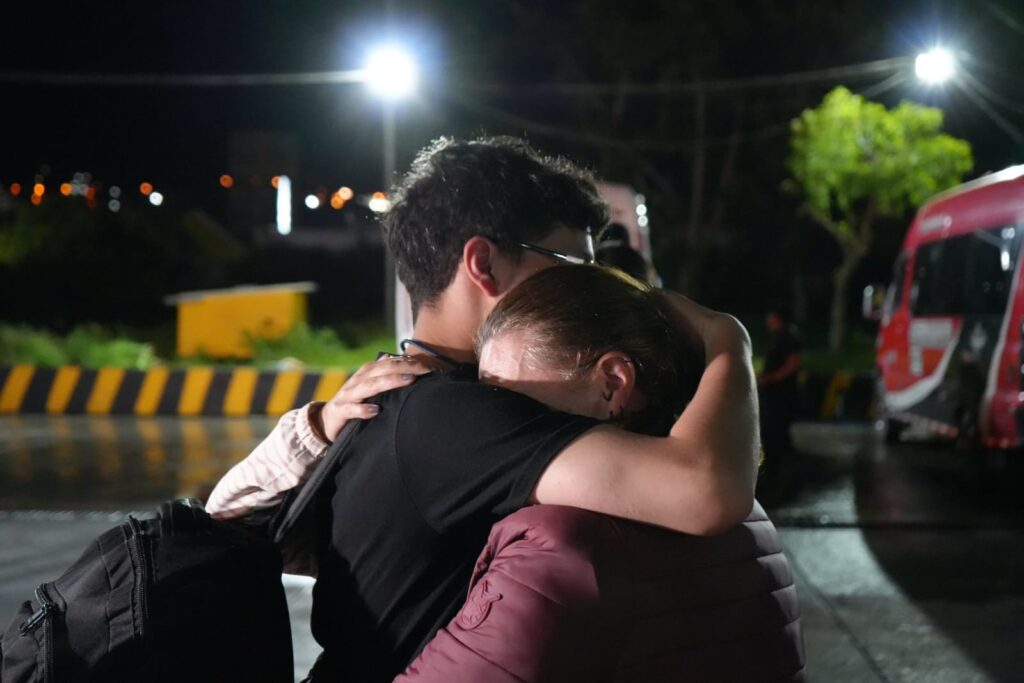 Madre e hijo se abrazan fuertemente en la Terminal de Transporte de Pasto durante la noche, tras encontrarse nuevamente en Nariño luego de un operativo de retorno seguro desde el Cauca. Uno de ellos lleva un morral negro y el otro una chaqueta acolchada color vino tinto. Al fondo se observan luces y un bus rojo.