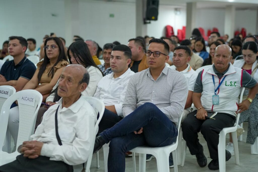 Asistentes sentados en sillas blancas durante una audiencia pública en Sandoná. Dos sillas al frente tienen etiquetas que dicen "ALCALDE". Una persona a la derecha lleva un chaleco con las letras "L.S.C".