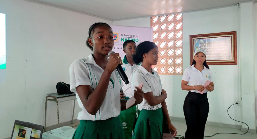 Una estudiante del Instituto Técnico Industrial Nacional (ITIN) en Tumaco habla por micrófono durante una jornada de sensibilización de la Estrategia Integral de Prevención de Delitos en Niños, Niñas y Adolescentes. Al fondo se observa el logo de la Gobernación de Nariño y otros compañeros participando.
