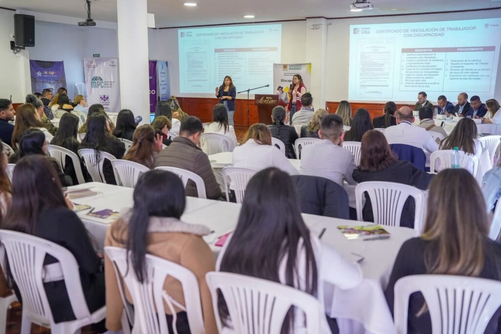 Reunión presencial en un auditorio lleno, con dos mujeres presentando en un podio al fondo sobre la vinculación laboral de personas con discapacidad, mientras los asistentes escuchan desde sus mesas.