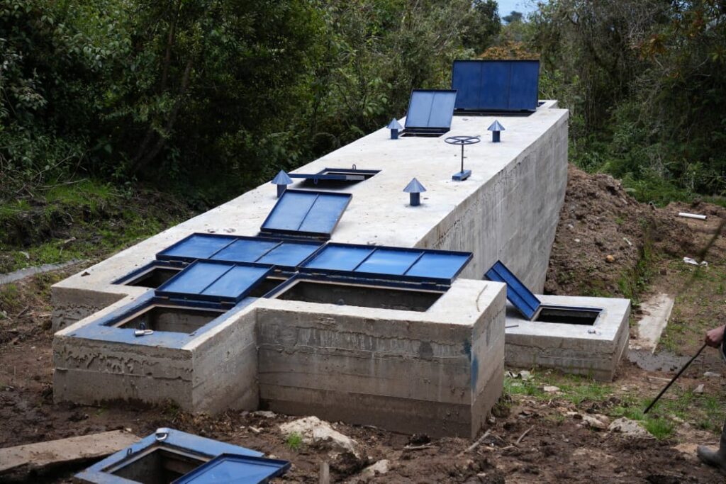 Estructura de concreto de un tanque desarenador con tapas metálicas azules, rodeada de vegetación, como parte del sistema de acueducto rural en Nariño.