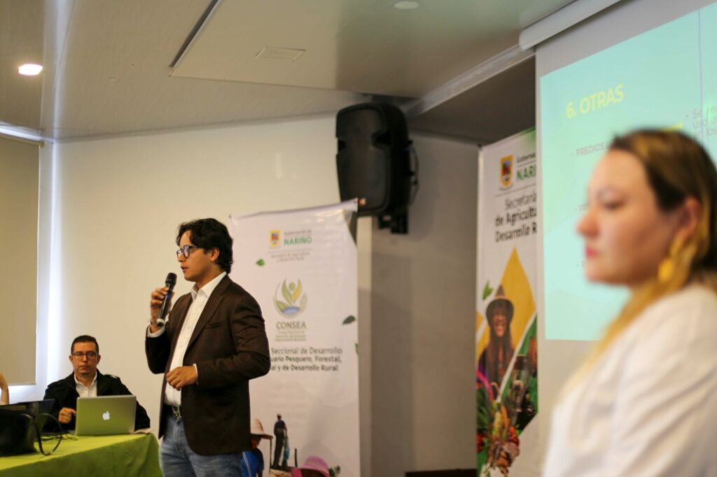 Andrés Acosta, funcionario de la Secretaría de Agricultura Departamental, interviene en la sesión del CONSEA junto a un delegado de la Agencia Nacional de Tierras durante la explicación técnica de la ruta de formalización de predios. Al fondo se observan pendones institucionales de la Gobernación de Nariño y el CONSEA.