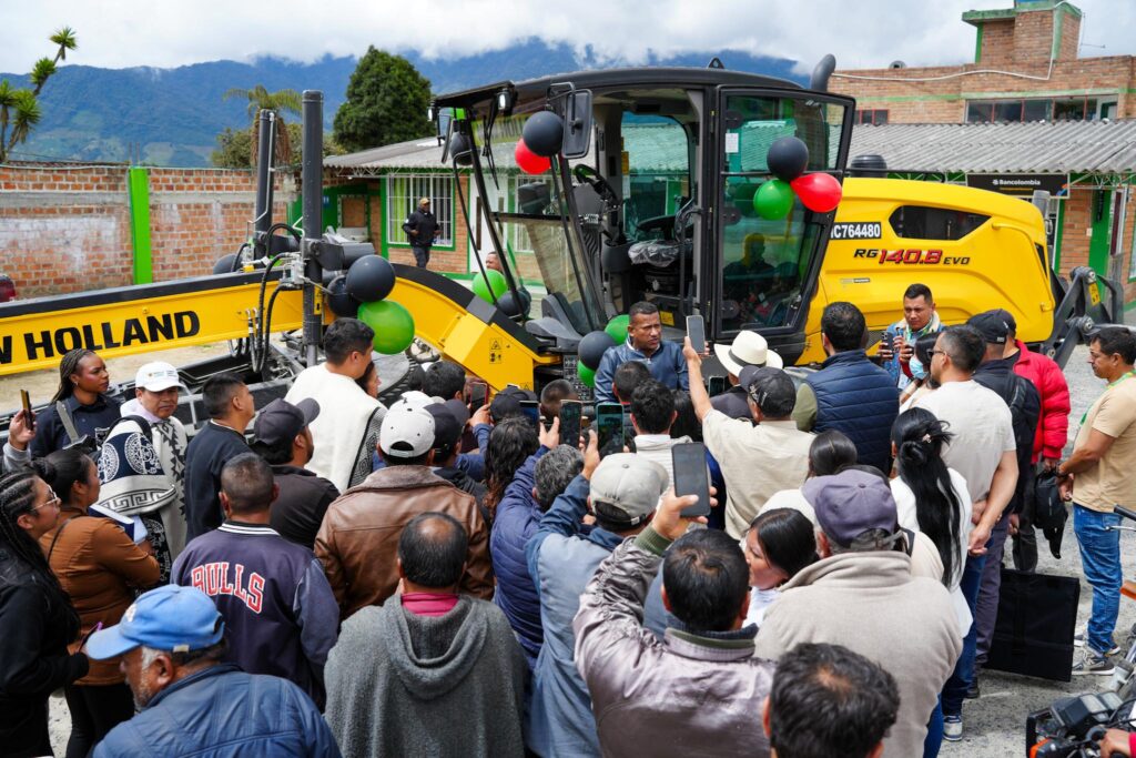 Gobernador de Nariño, Luis Alfonso Escobar Jaramillo, hablando a la comunidad junto a una motoniveladora amarilla New Holland RG140.B Evo decorada con globos rojos, verdes y negros. Varias personas están tomando fotos con teléfonos celulares.