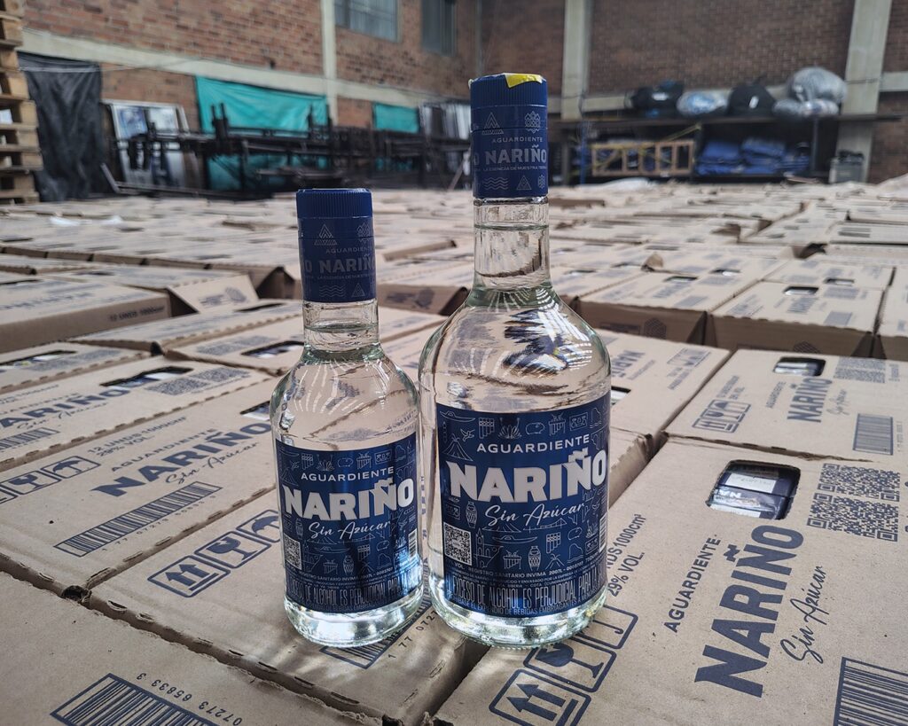 Dos botellas de Aguardiente Nariño Azul Sin Azúcar, en presentaciones de 750cc y 375cc, dispuestas en primer plano sobre cajas de cartón de la marca dentro de una bodega de la Gobernación de Nariño. Las botellas exhiben su etiqueta azul característica y sellos de seguridad.