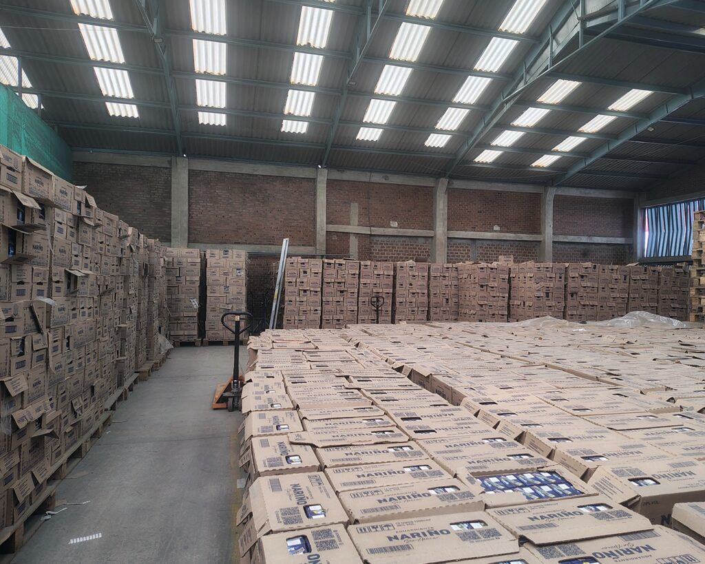 Bodega de almacenamiento de la Gobernación de Nariño en Pasto, repleta de cajas apiladas de Aguardiente Nariño listas para su distribución comercial. El almacén de estructura industrial muestra cientos de cajas del licor insignia del departamento, organizadas para el proceso de venta de 2026 anunciado por la Subsecretaria de Rentas. Un carro manual de carga es visible entre las estibas.