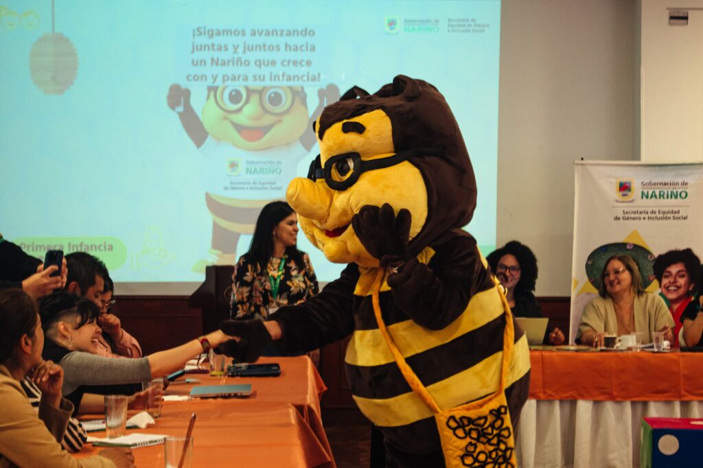 Beecky, la abeja protectora de la Gobernación de Nariño, saluda a una asistente durante la socialización del cronograma de la Brigada Protectora 2026. Al fondo, una proyección con el mensaje 'Sigamos avanzando juntas y juntos hacia un Nariño que crece con y para su infancia'.
