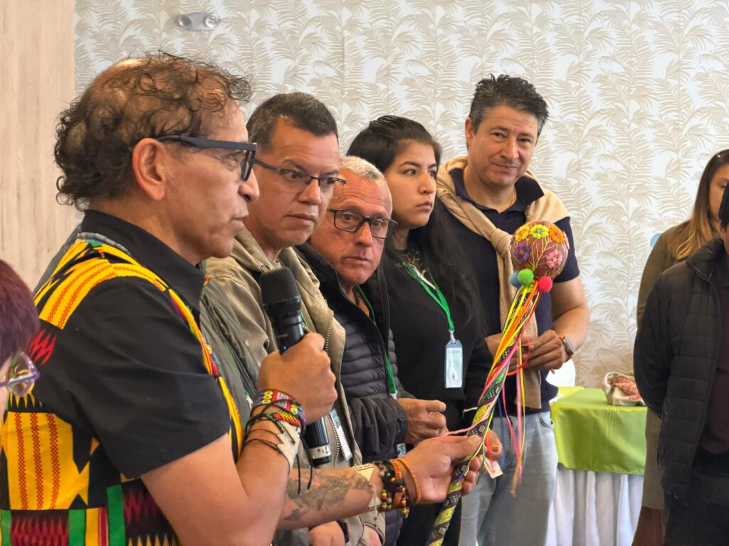 El director Administrativo de Cultura de Nariño, Álvaro Reyes, habla por micrófono durante la sesión del Consejo Departamental de Cultura. Viste un chaleco con patrones tradicionales coloridos y sostiene un bastón artesanal decorado. Al fondo, consejeros y asistentes escuchan atentos.
