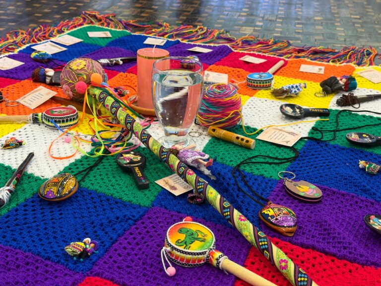 Altar o instalación simbólica sobre una manta tejida de cuadros multicolores en el suelo, con artesanías de Nariño, un bastón de mando decorado, instrumentos musicales pequeños, un vaso de agua, una vela y notas escritas, usada en la sesión del Consejo Departamental de Cultura.