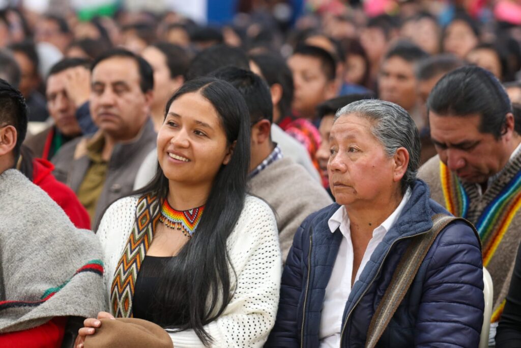 Mujer joven con collar artesanal indígena sonríe junto a una mujer mayor y una multitud durante el encuentro étnico en el municipio de Aldana, Nariño.