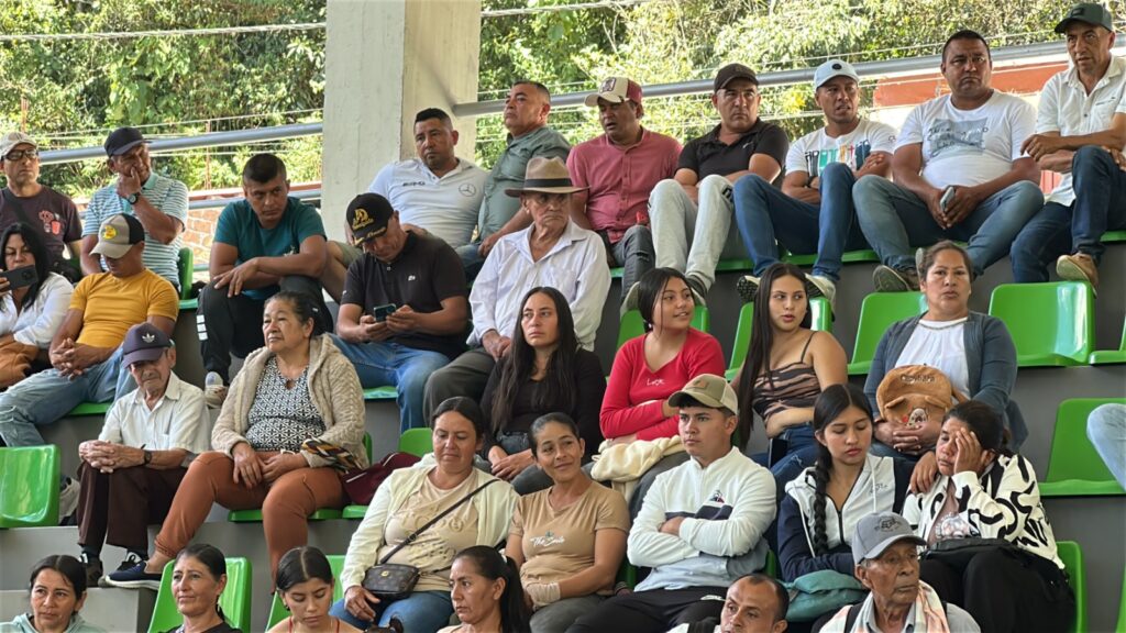 Un grupo numeroso de personas, incluyendo adultos mayores, mujeres, jóvenes y hombres campesinos, se encuentran sentados juntos en las graderías con sillas plásticas verdes de un coliseo cubierto. Sus expresiones denotan preocupación y atención.