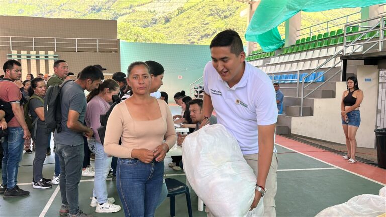 El Secretario de Paz y Convivencia Ciudadana de la Gobernación de Nariño, Álex González, con camiseta tipo polo blanca institucional y pantalón claro, sonríe mientras entrega un gran bulto blanco de ayuda humanitaria a una mujer de cabello recogido y blusa color beige. Al fondo se observa una fila de personas en una cancha de coliseo y montañas verdes.