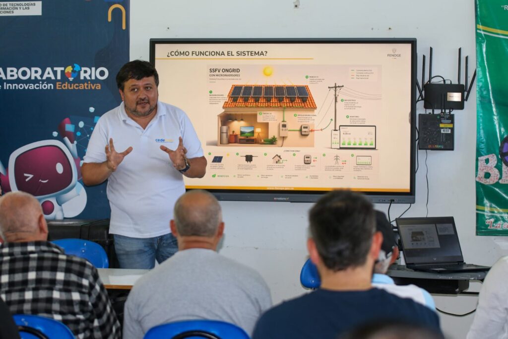 Un expositor de Cedenar, con camisa polo blanca y jeans, gesticula con las manos al socializar un sistema fotovoltaico frente a un público en Sandoná. Detrás de él, una pantalla digital titula ¿CÓMO FUNCIONA EL SISTEMA? y muestra un esquema técnico residencial de FENOGE llamado SSFV ONGRID con paneles solares