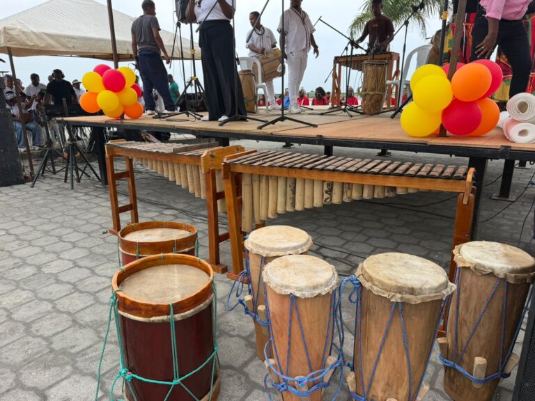 Kit de instrumentos musicales tradicionales expuestos en primer plano, que incluye dos marimbas de chonta, bombos y cununos, con un escenario de fondo en Tumaco.