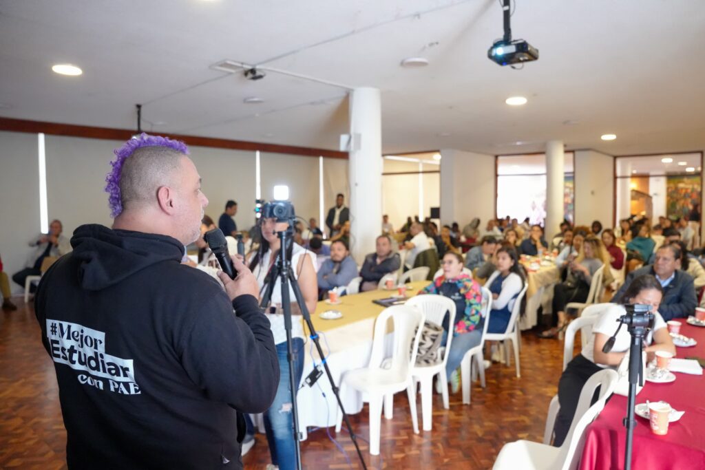 Vista desde atrás de un hombre de perfil con cabello teñido de morado y buzo negro que dice en la espalda "#Mejor Estudiar con PAE", quien habla por micrófono ante un auditorio lleno. El público se encuentra sentado en mesas organizadas dentro de un salón social amplio con equipos de filmación en primer plano.
