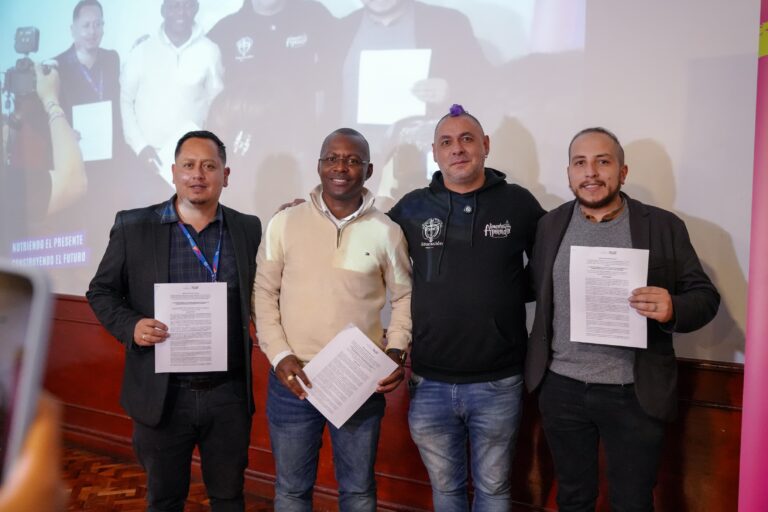 Cuatro hombres posan sonrientes sosteniendo hojas oficiales del programa de alimentación escolar. El segundo de izquierda a derecha es un funcionario afrodescendiente con saco color beige y camisa blanca; a su lado derecho hay un hombre con buzo negro estampado y cabello teñido de morado en estilo cresta, y los extremos laterales están ocupados por dos hombres de chaqueta oscura. Al fondo se proyecta la misma imagen en una pantalla grande.