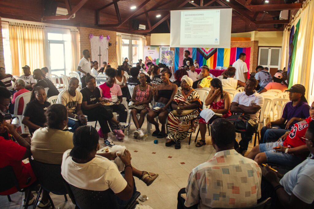 Líderes y organizaciones de la población afro LGBTIQ+ víctimas del conflicto armado reunidos en círculo durante una mesa de trabajo en Tumaco, bajo banderas de la diversidad y una pantalla de proyecciones.