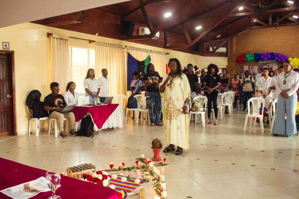 Una lideresa afrodescendiente interviene con micrófono en mano durante el encuentro "Diálogos para la inclusión" en Tumaco. Al frente se observa un altar simbólico con la bandera LGBTIQ+, velas, flores y una marimba.