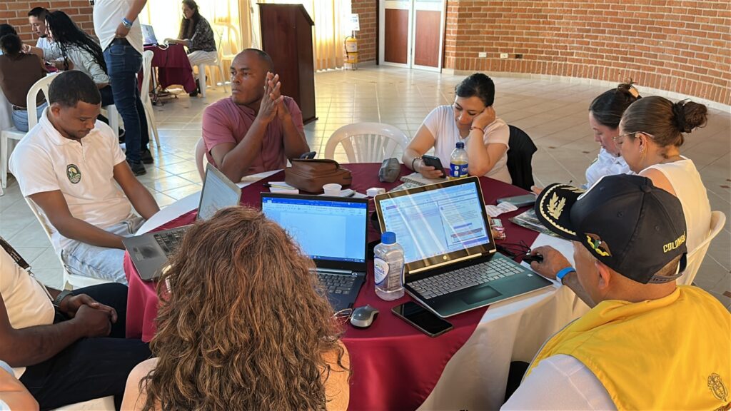 Grupo de funcionarios y representantes comunitarios reunidos alrededor de una mesa circular con computadores portátiles, trabajando en la aprobación de planes operativos en Tumaco.