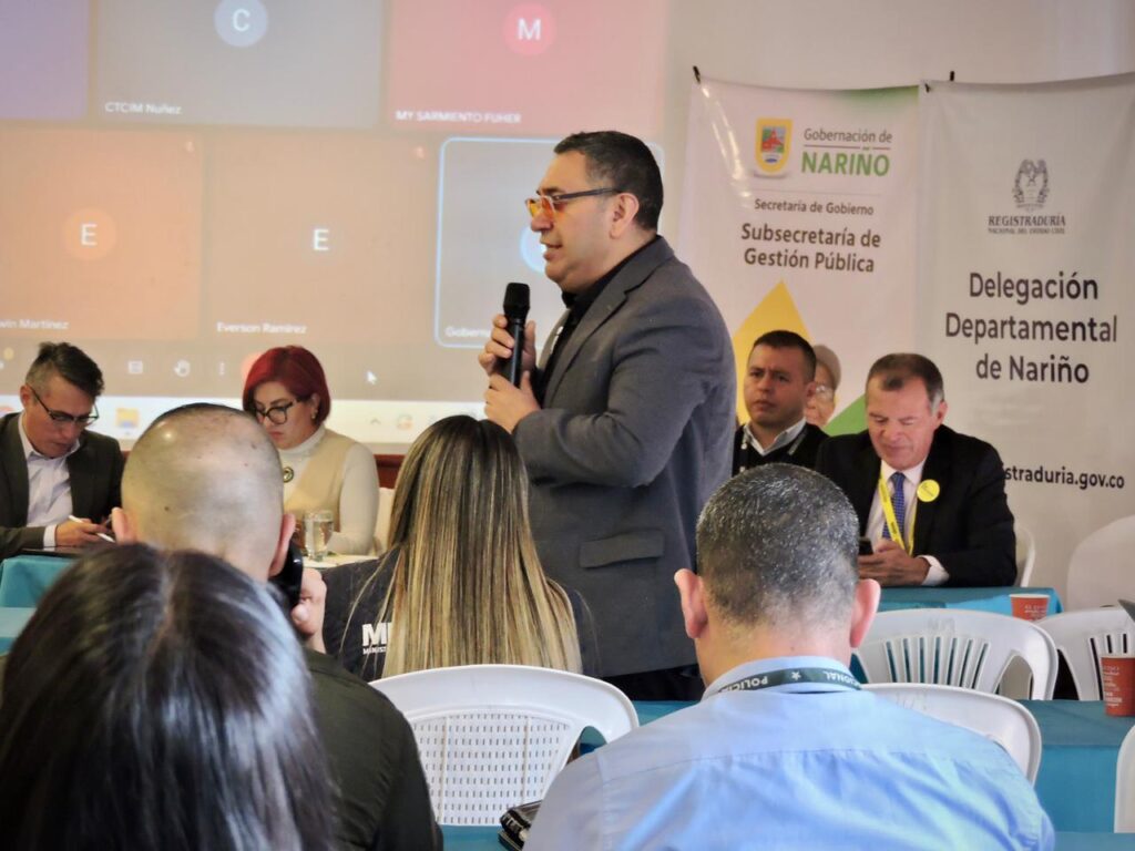 Un funcionario interviene en la Comisión de Coordinación y Seguimiento Electoral en Nariño. Viste saco gris, camisa negra, gafas y sostiene un micrófono con la mano derecha, mientras que detrás de él hay una pantalla con asistentes virtuales y banners institucionales de la Gobernación de Nariño y la Registraduría.