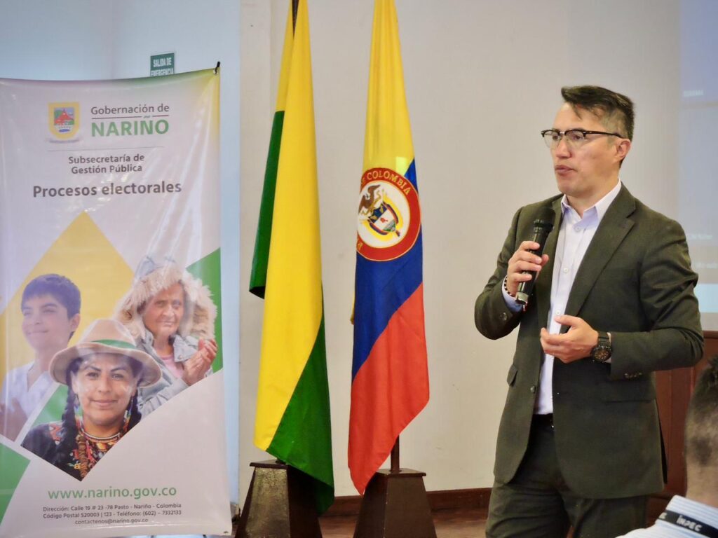 Fredy Andrés Gámez Castrillón, Secretario de Gobierno Departamental de Nariño, interviene en la Comisión de Coordinación y Seguimiento Electoral. Viste traje formal verde oliva, camisa blanca y gafas, sosteniendo un micrófono con su mano derecha. A su lado se observan las banderas de Colombia y del departamento de Nariño, junto a un pendón de la Subsecretaría de Gestión Pública sobre procesos electorales.