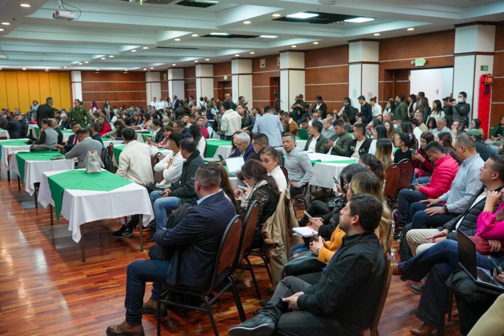 Un auditorio amplio y concurrido con mesas rectangulares vestidas con manteles blancos y verdes. Numerosos ciudadanos, líderes y expertos se encuentran sentados prestando atención al evento, mientras algunos revisan apuntes y computadores portátiles.
