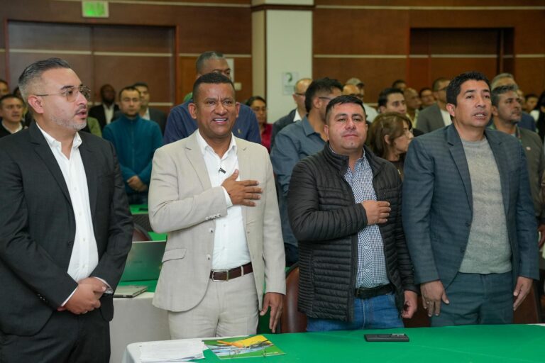 El Gobernador de Nariño, Luis Alfonso Escobar Jaramillo, de traje formal color beige y camisa blanca, se encuentra de pie con la mano derecha sobre el pecho durante los actos protocolares. Está acompañado en la mesa principal por otros tres funcionarios y asistentes al fondo.