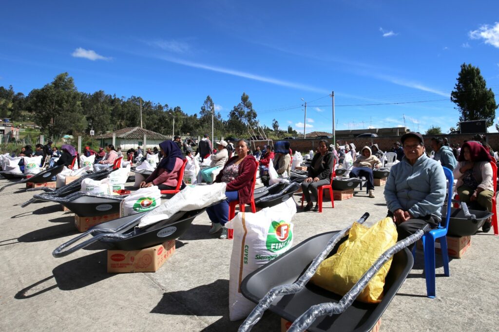 Familias campesinas e indígenas de Ipiales sentadas en una plaza comunitaria durante una jornada de entregas agropecuarias. En primer plano, se observan carretillas tipo bugui de color negro marca Herragro y bultos de alimento concentrado para cuyes bajo un cielo despejado.