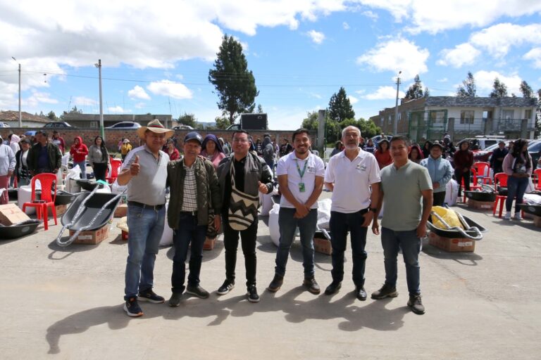 Un grupo de seis hombres, entre ellos técnicos y líderes del proyecto agropecuario, posan sonrientes en una plaza comunitaria al aire libre en Ipiales. Al fondo se observan familias beneficiarias, carretillas tipo bugui, bultos de insumos distribuidos y viviendas bajo un cielo parcialmente nublado.