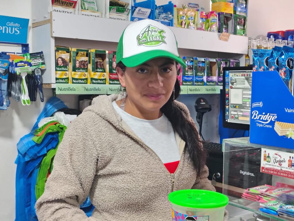Retrato en primer plano de Sara Marcela Chacua, propietaria de un granero en Ipiales, usando una gorra blanca y verde con el logotipo de la campaña "Aquí Todo es Legal" de la Gobernación de Nariño, ubicada detrás del mostrador de su establecimiento comercial.