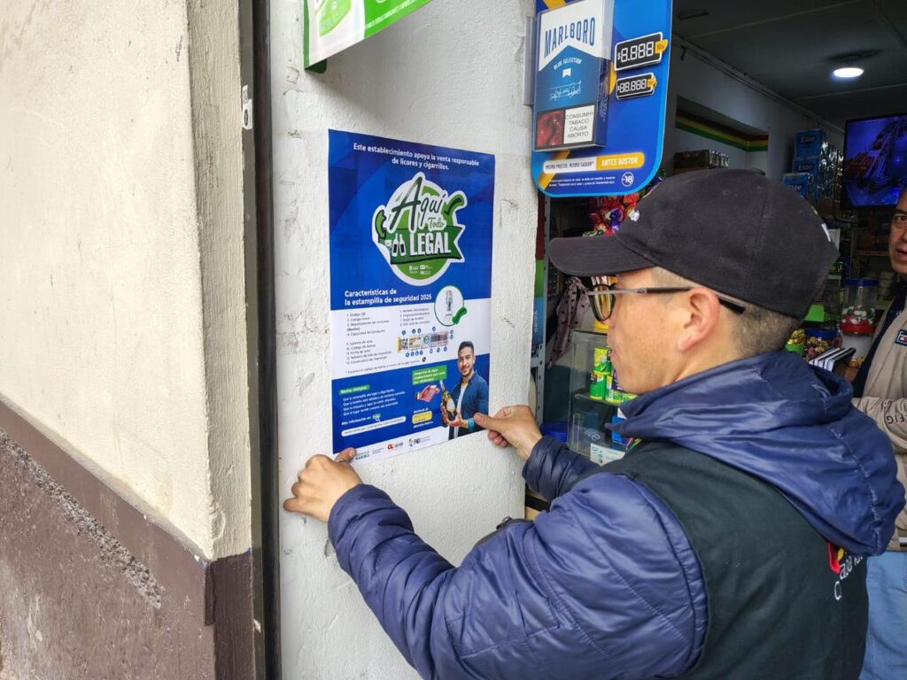 Funcionario del Grupo Operativo Anticontrabando de la Gobernación de Nariño de perfil, pegando un afiche informativo de la campaña "Aquí Todo es Legal" sobre las características de la estampilla de seguridad en la pared exterior de un local comercial.