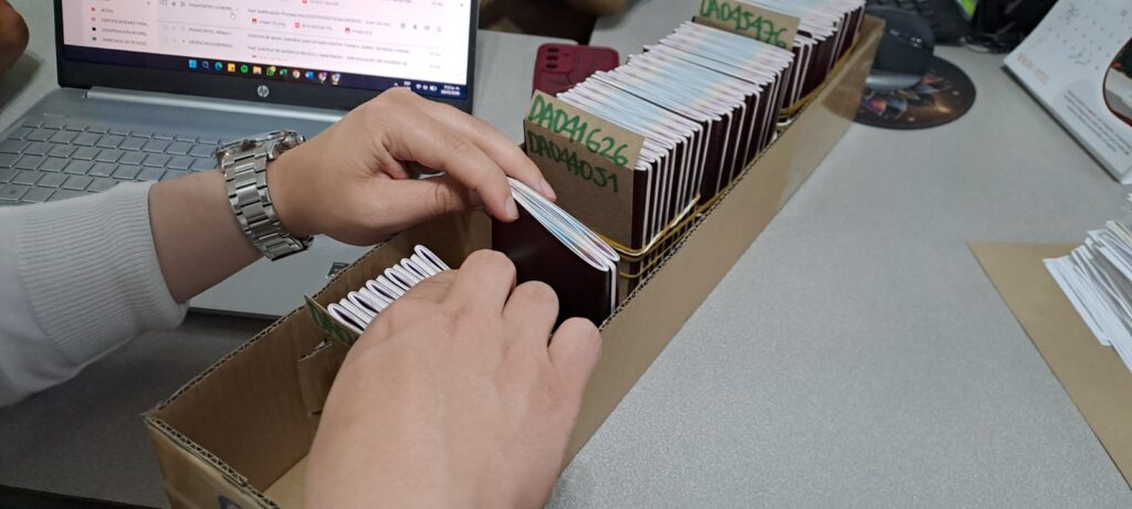 Manos de una funcionaria organizando y revisando lotes del nuevo pasaporte colombiano ordenados en cajas sobre un escritorio junto a un computador portátil en la Oficina de Pasaportes de Nariño.
