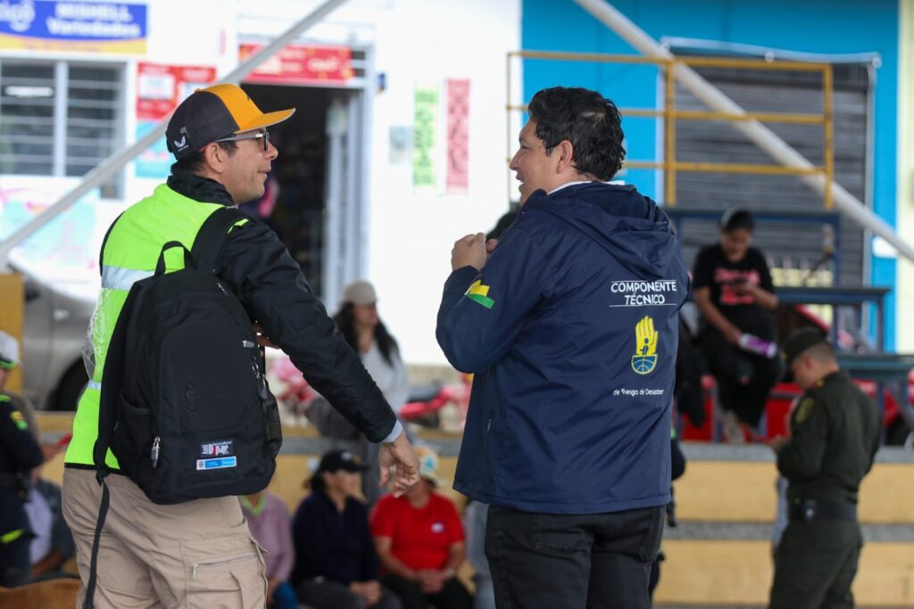 Dos funcionarios públicos conversando durante el simulacro de erupción volcánica. Uno viste chaleco reflectivo verde y morral negro; el otro lleva una chaqueta azul marino institucional con la inscripción "Componente Técnico de Riesgo de Desastres".