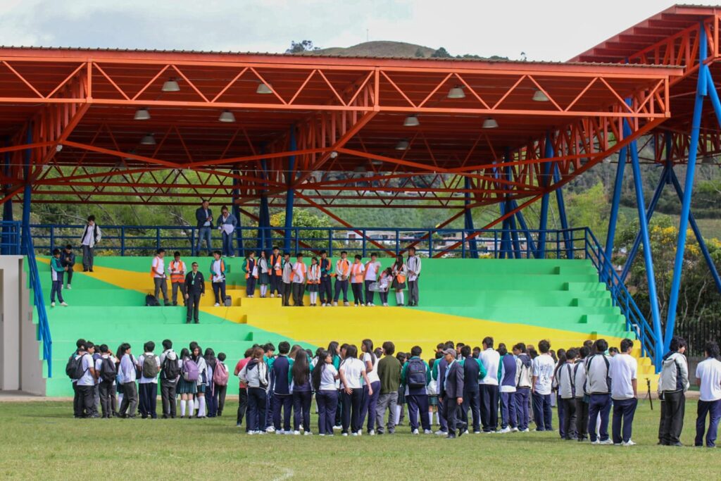 Estudiantes de una institución educativa pública concentrados en una cancha de fútbol junto a una tribuna con techado naranja, siguiendo las indicaciones de los brigadistas escolares durante el simulacro de evacuación.