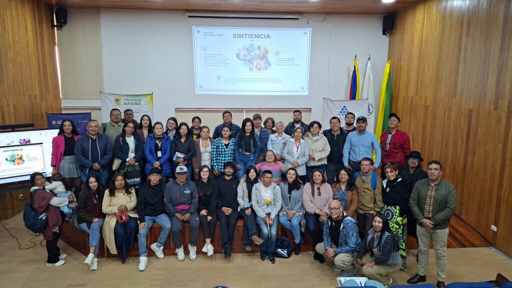 Un grupo grande de personas posa para una foto grupal en un escenario, con una pantalla de proyección que dice "SINTIENCIA" detrás de ellos.Título: Foto Grupal de Capacitación.
