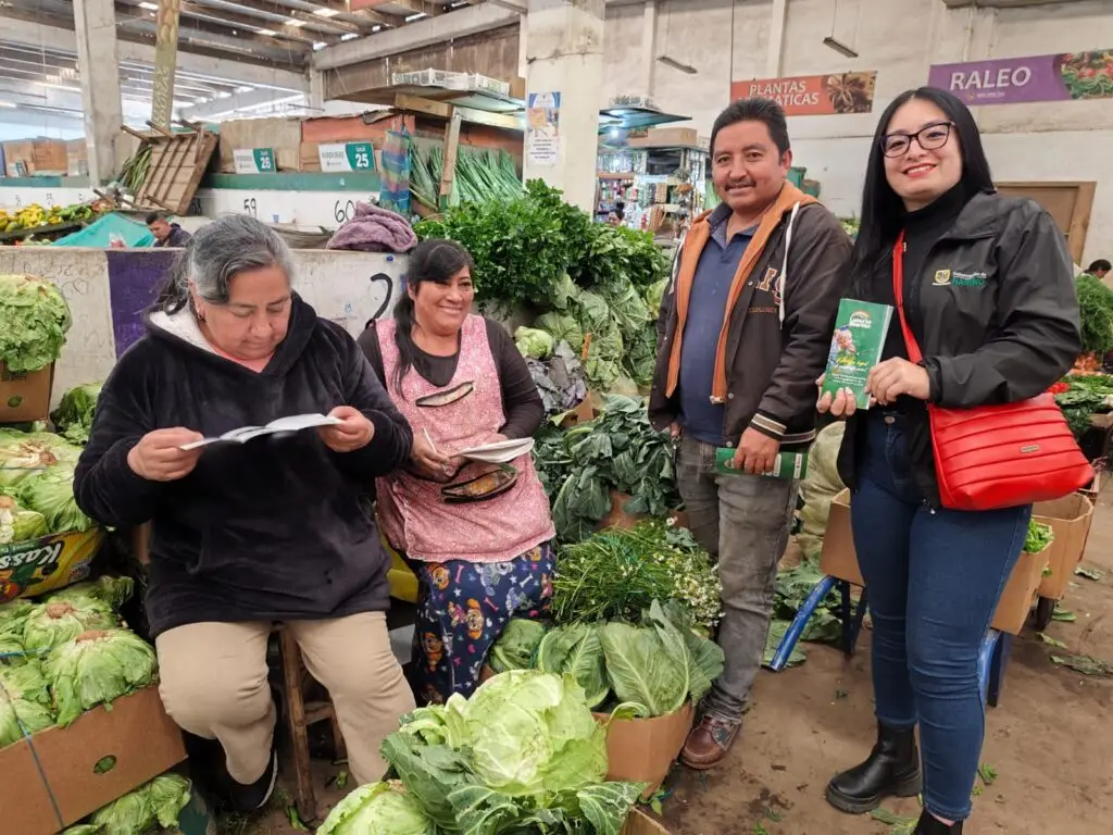 Una funcionaria de la Gobernación de Nariño entrega folletos informativos de la campaña "Juega Legal" de la Lotería de Nariño a vendedores de verduras en una plaza de mercado.