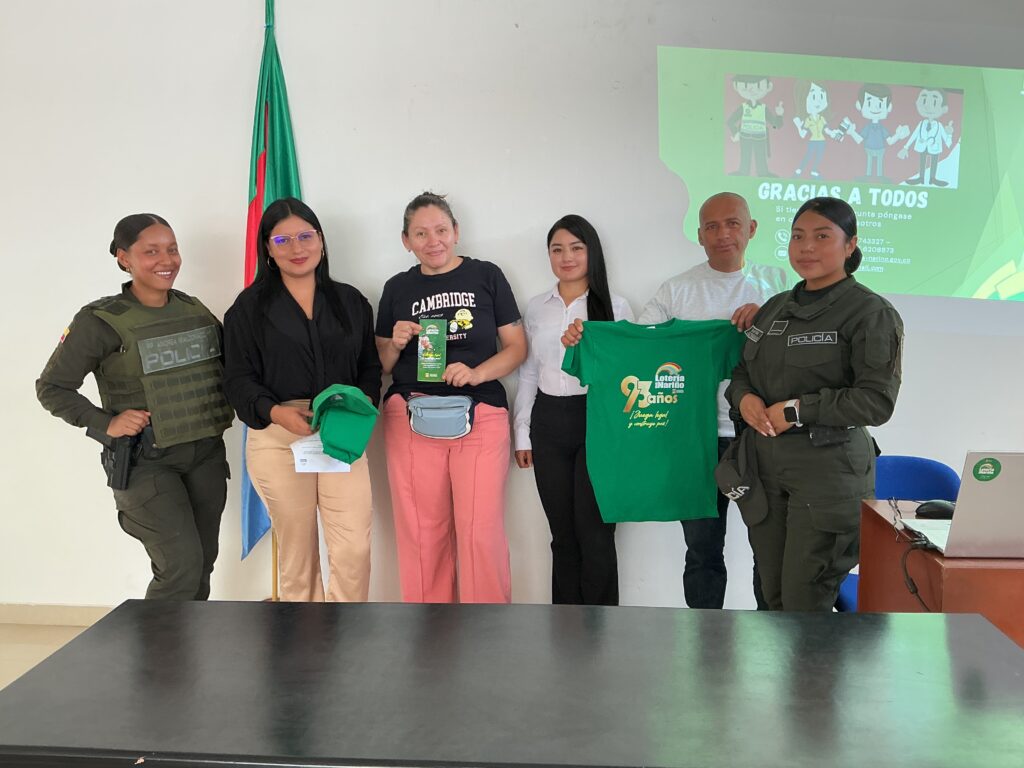Integrantes de la Policía Nacional, funcionarios de la Lotería de Nariño y ciudadanos posan sosteniendo una camiseta verde de los 93 años de la entidad con el lema "¡Juega legal y construye paz!" en una capacitación.