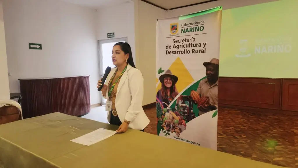 Funcionaria de la Secretaría de Agricultura y Desarrollo Rural de Nariño exponiendo con micrófono al lado del pendón oficial de la entidad durante la asamblea del Comité Acuícola.