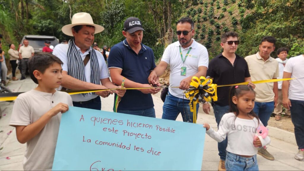 Acto de inauguración de la vía en San Pablo, Nariño. El alcalde Mario Trujillo con gorra que dice 'ALCALDE', junto a funcionarios y miembros de la comunidad, cortan una cinta amarilla y negra sobre la nueva placa huella. Al frente, dos niños sostienen una pancarta azul de agradecimiento.