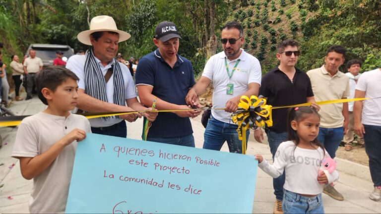 Acto de inauguración de la vía en San Pablo, Nariño. El alcalde Mario Trujillo con gorra que dice 'ALCALDE', junto a funcionarios y miembros de la comunidad, cortan una cinta amarilla y negra sobre la nueva placa huella. Al frente, dos niños sostienen una pancarta azul de agradecimiento.