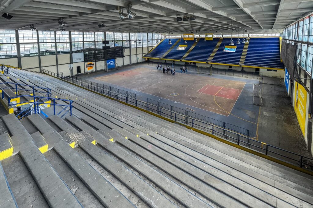 Vista interior panorámica del Coliseo Cubierto Sergio Antonio Ruano en Pasto, mostrando graderías de cemento vacías, la cancha de juego desgastada y un grupo de técnicos realizando inspección en el centro