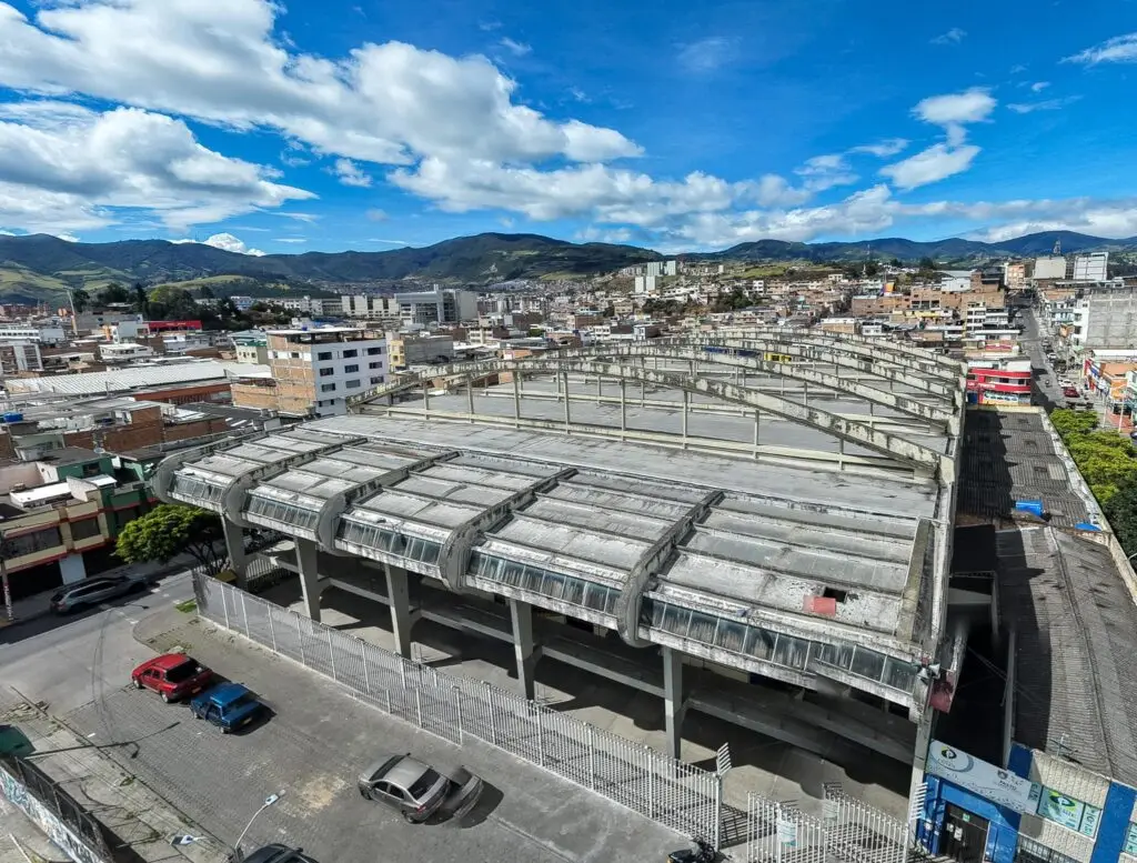 Vista aérea exterior del Coliseo Sergio Antonio Ruano en Pasto, mostrando su distintivo techo abovedado de concreto en mal estado, rodeado por la ciudad y montañas bajo un cielo parcialmente nublado.