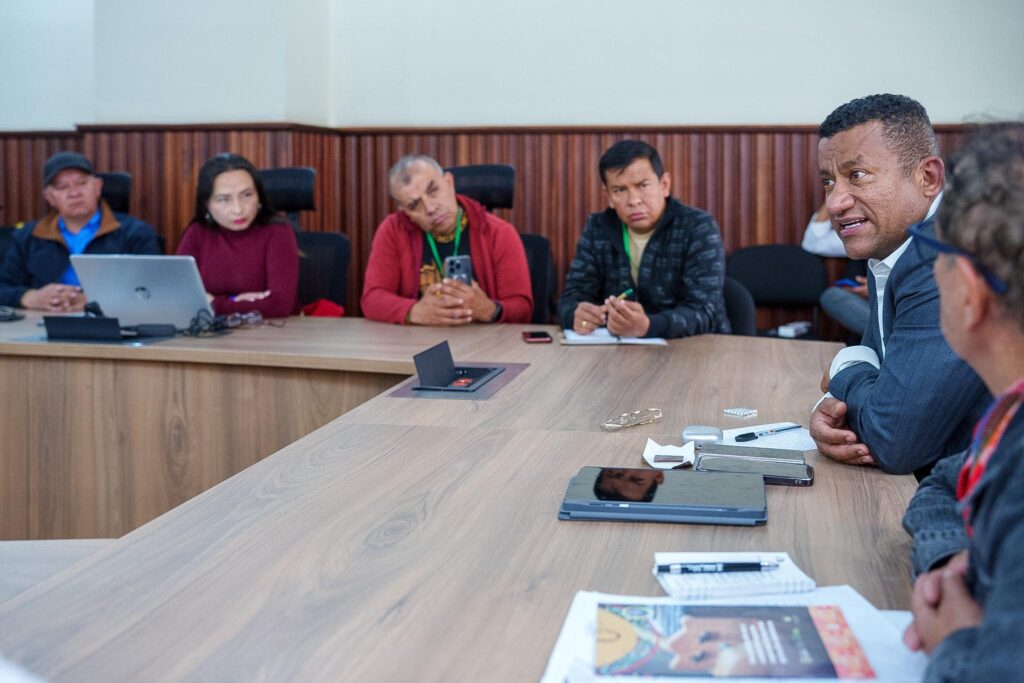 El Gobernador de Nariño, Luis Alfonso Escobar Jaramillo, habla de perfil en primer plano mientras los integrantes del Consejo Departamental de Cultura lo escuchan atentos alrededor de una mesa de juntas.