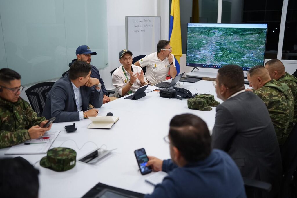 Integrantes del Puesto de Mando Unificado (PMU) sentados alrededor de una mesa blanca de juntas analizan un mapa satelital de la Cordillera de Nariño proyectado en un televisor grande al fondo.