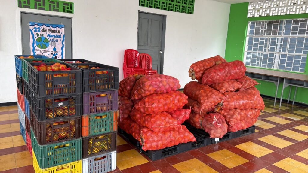 Alimentos del PAE apilados en un salón de clases: canastillas plásticas de colores con frutas frescas y bultos de color rojo con productos agrícolas sobre estibas de madera.
