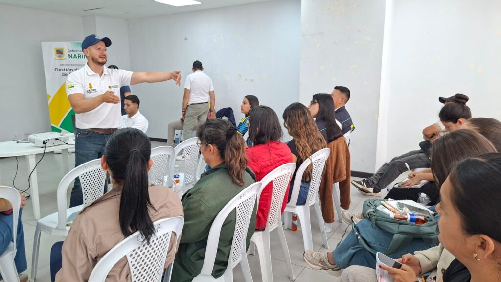 Vista de perfil del profesional Jorge Tovar dictando un taller técnico de Gestión del Riesgo de Nariño a representantes de los Consejos Municipales sentados en hileras. Al fondo se aprécian pendones informativos de la Gobernación.