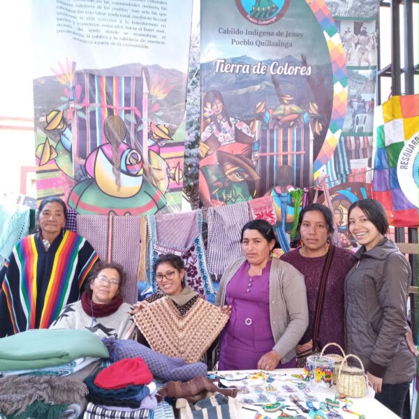 Grupo de cinco mujeres artesanas del Cabildo Indígena de Jenoy, Pueblo Quillasinga, posando sonrientes detrás de una mesa de exhibición de textiles tradicionales que incluye ruanas, bufandas, bolsos y artesanías en un evento institucional.