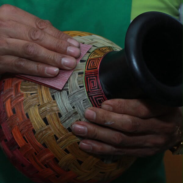 Primer plano de las manos de un artesano nariñense aplicando una lija a la superficie de un jarrón de madera decorado con la técnica ancestral del Barniz de Pasto. El jarrón presenta diseños geométricos con degradados de color rojo, naranja y amarillo en su cuerpo, y un cuello negro pulido con finas líneas tradicionales.