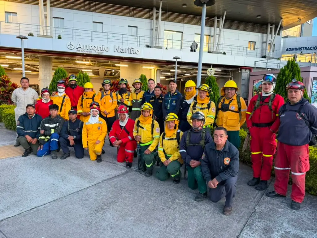 Fotografía grupal de unos veinticinco bomberos con uniformes de fatiga y trajes de protección amarillos y rojos, algunos con cascos y equipo de montaña, posando frente a la entrada del Aeropuerto Antonio Nariño.