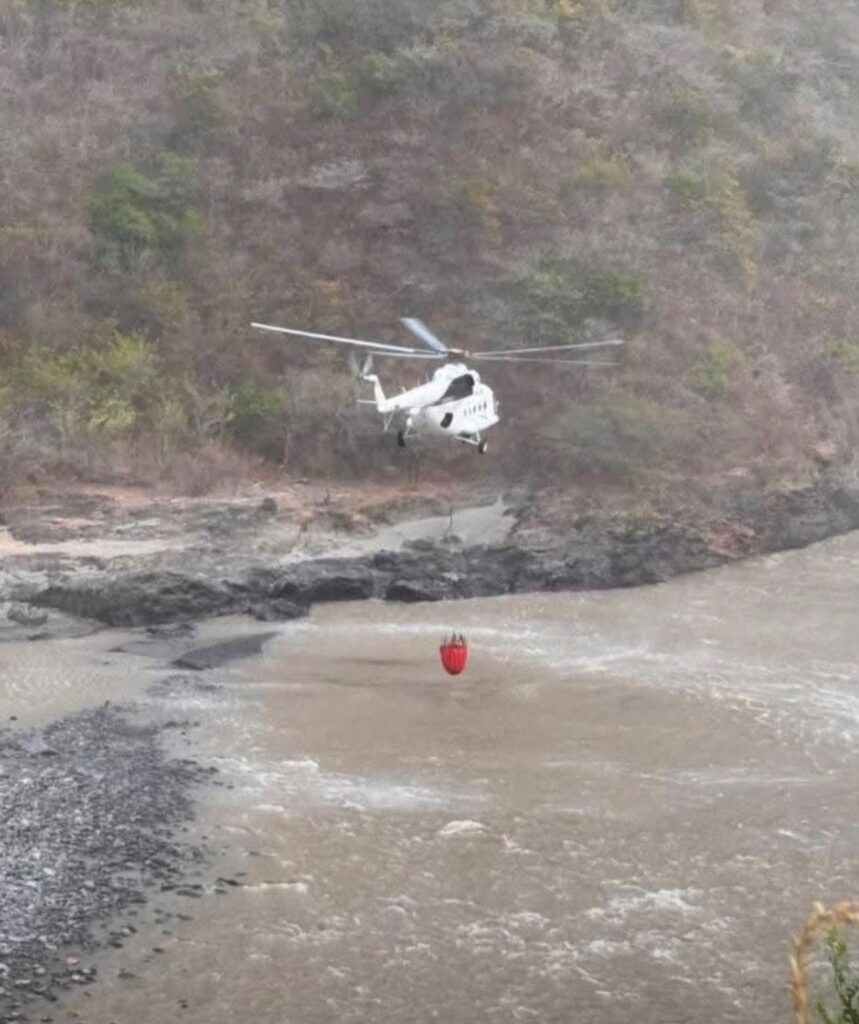 Un helicóptero blanco MI-17 vuela a baja altura sobre el cauce ancho del Río Patía, suspendiendo un gran cubo rojo de Bambi Bucket justo por encima del agua para abastecerse, frente a una ladera de montaña cubierta de vegetación seca.